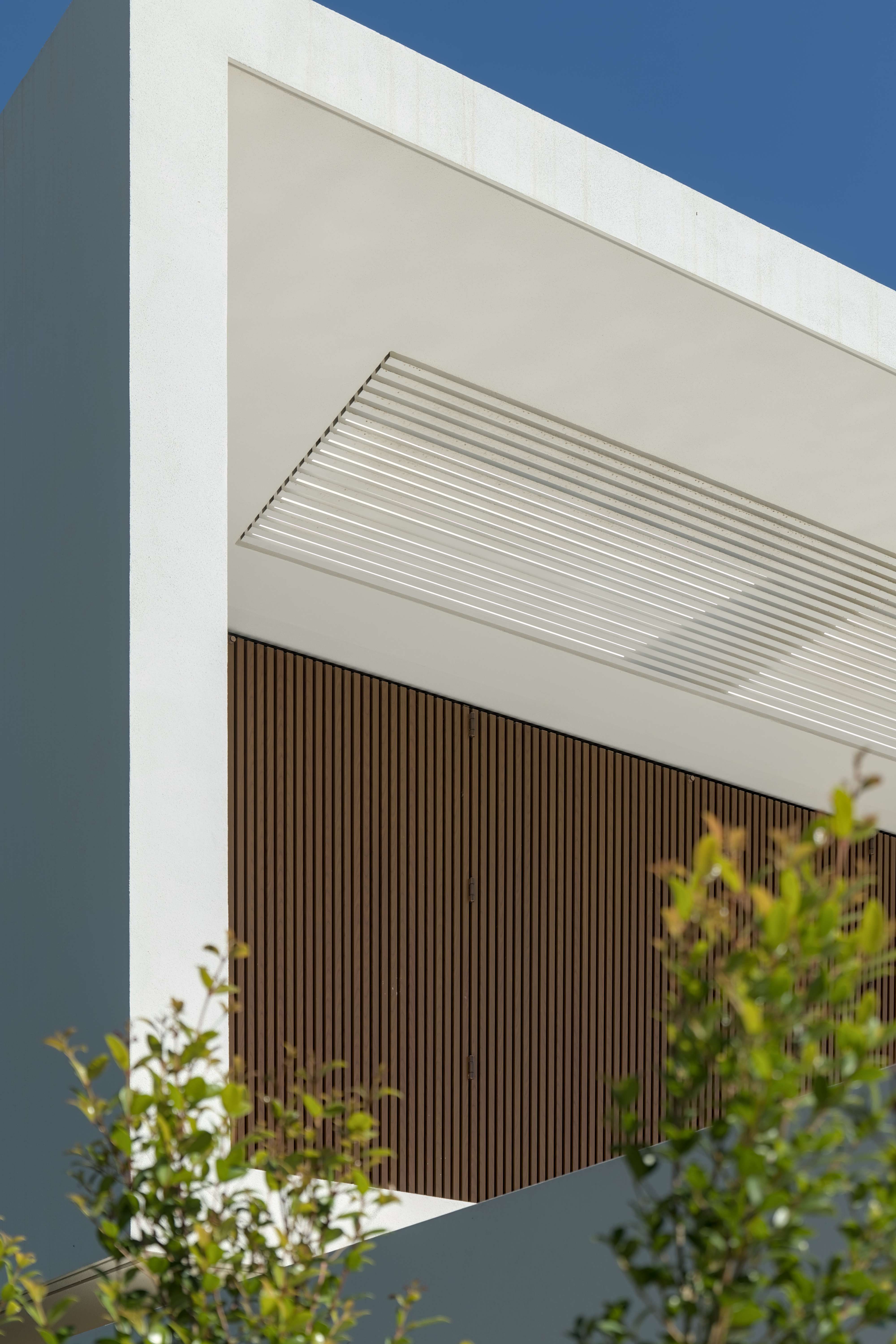 Pormenor da fachada de uma casa contemporânea onde se destaca um gradeamento metálico com textura de madeira. O enquadramento é emoldurado por duas árvores pequenas nos cantos inferiores da imagem.