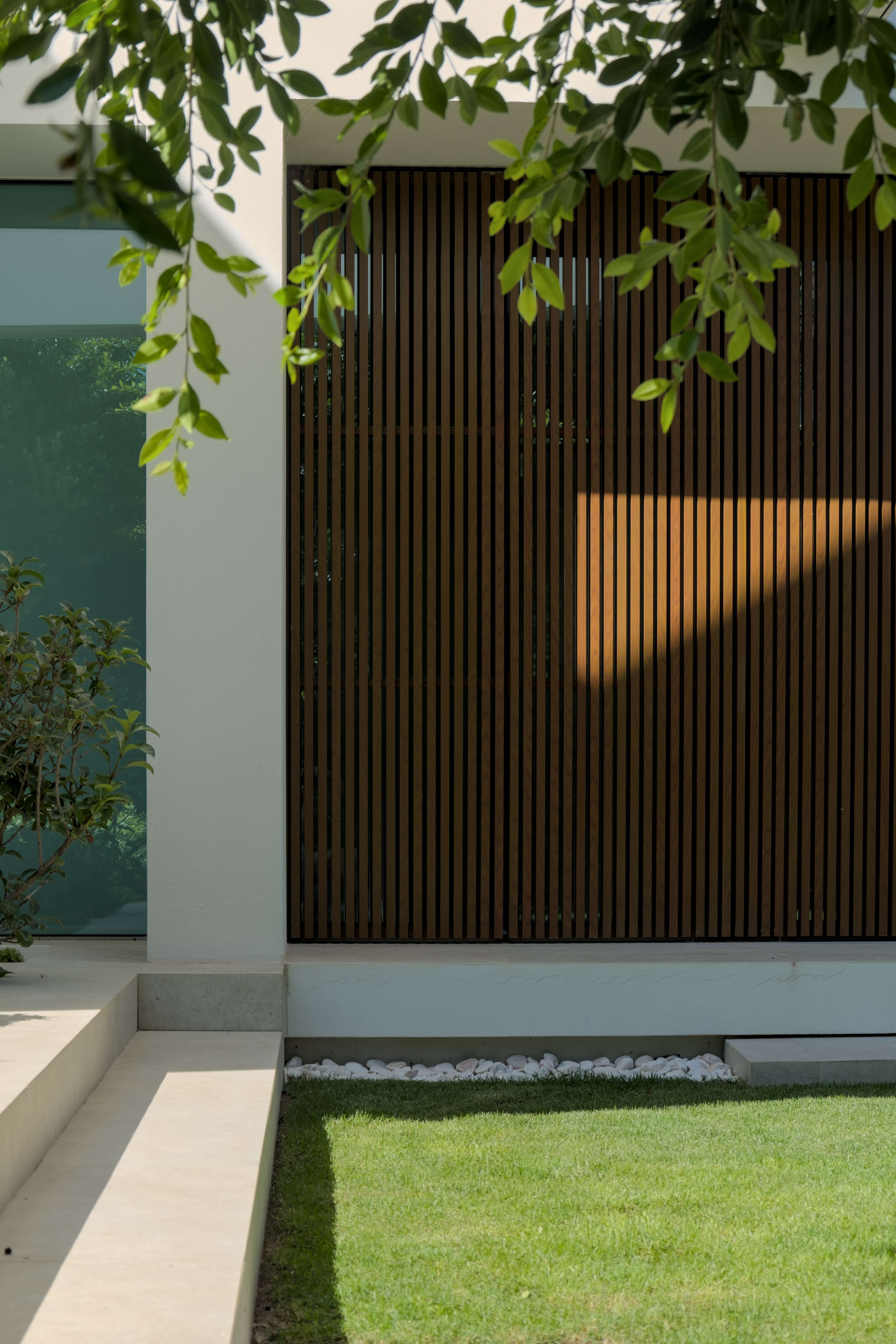 Jardim de uma casa contemporânea. A casa tem uma janela que se extende por todo o cumprimento e altura da parede e está protegida por um gradiamento metálico com um aspeto de madeira.