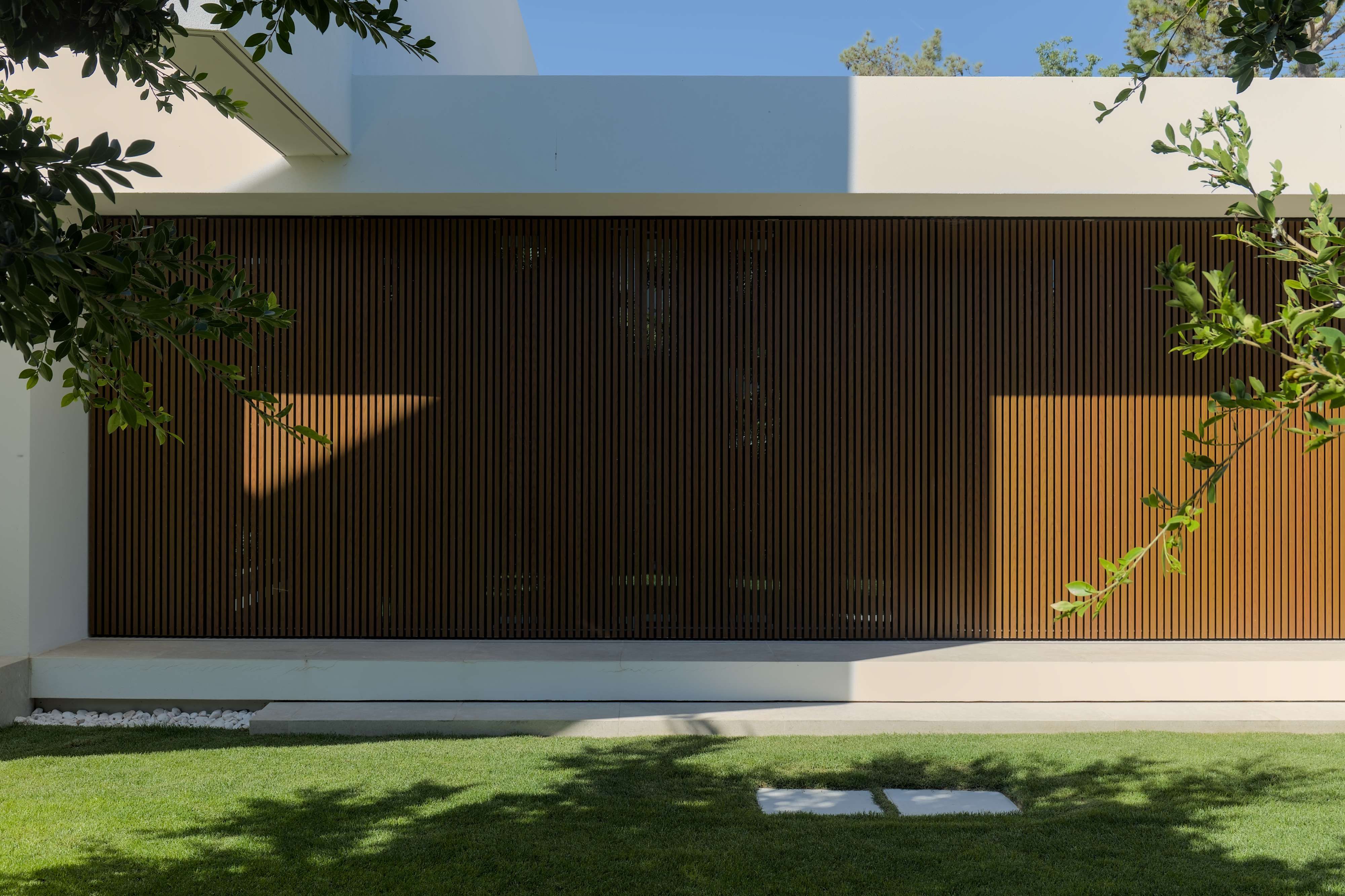 Jardim de uma casa contemporânea. A casa tem uma janela que se extende por todo o cumprimento e altura da parede e está protegida por um gradiamento metálico com um aspeto de madeira.