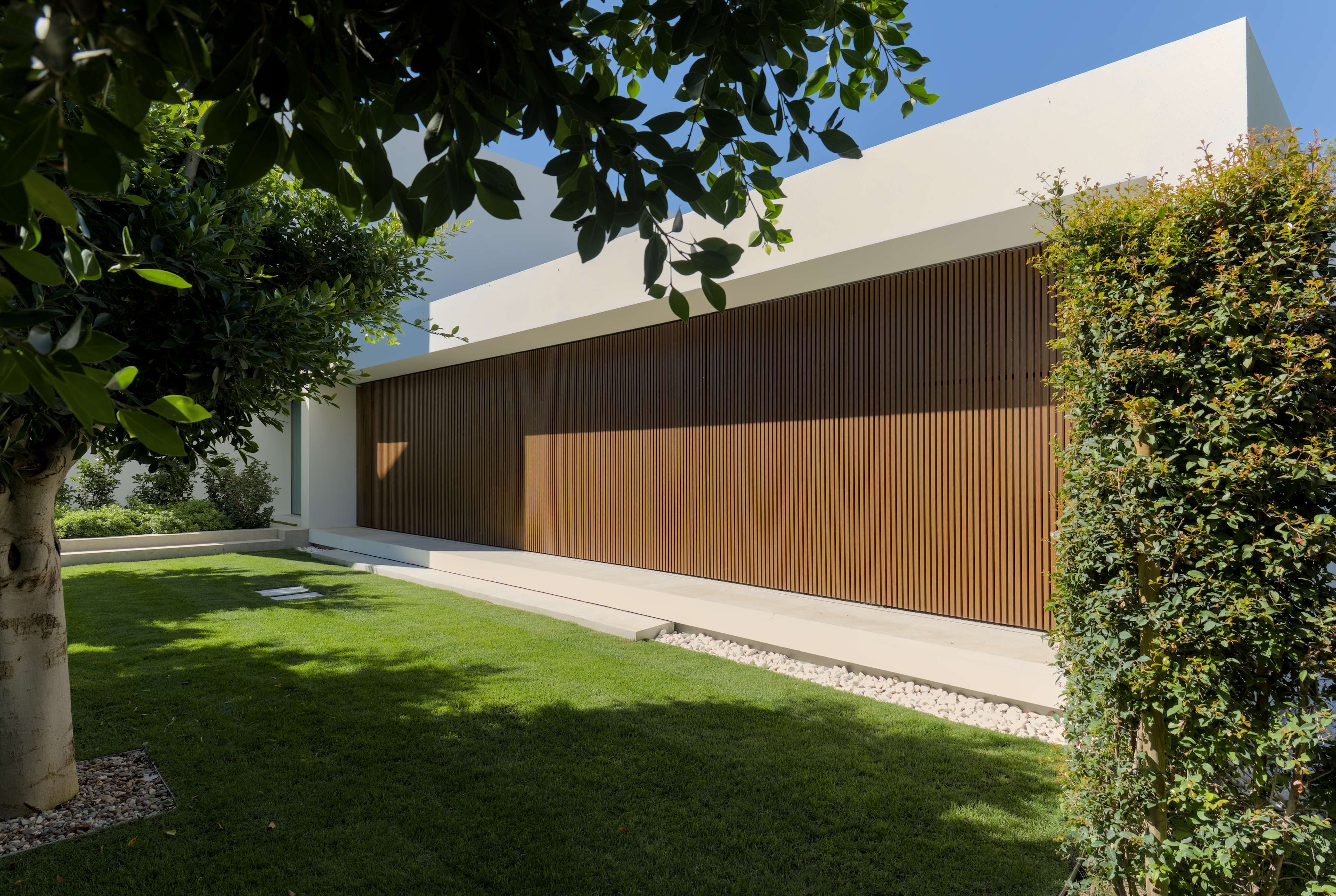 Jardim de uma casa contemporânea. A casa tem uma janela que se extende por todo o cumprimento e altura da parede e está protegida por um gradiamento metálico com um aspeto de madeira.