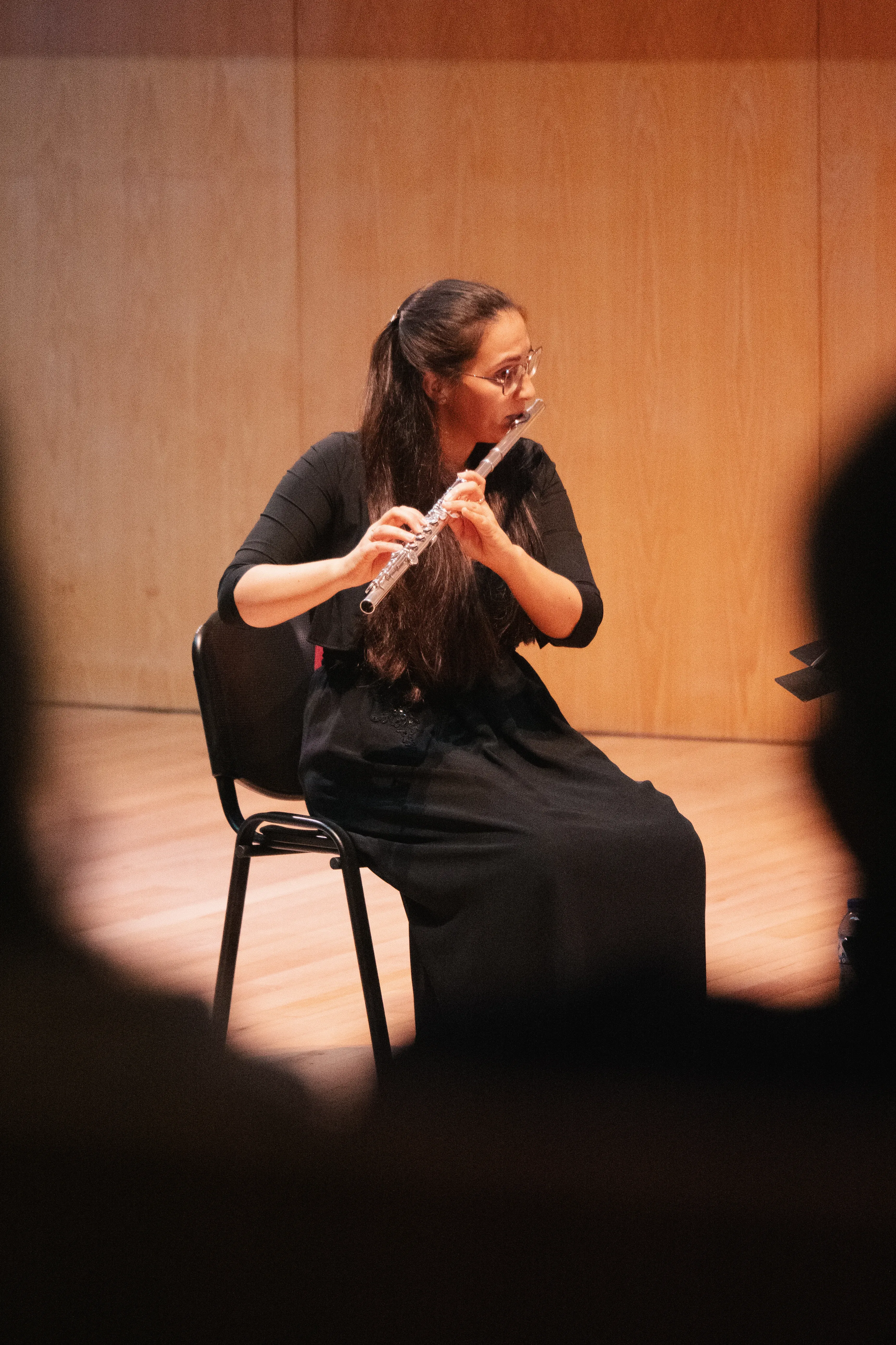 Uma artista toca flauta em palco. Em primeiro plano vê-se o perfil de alguns elementos da plateia que a emolduram no enquadramento.