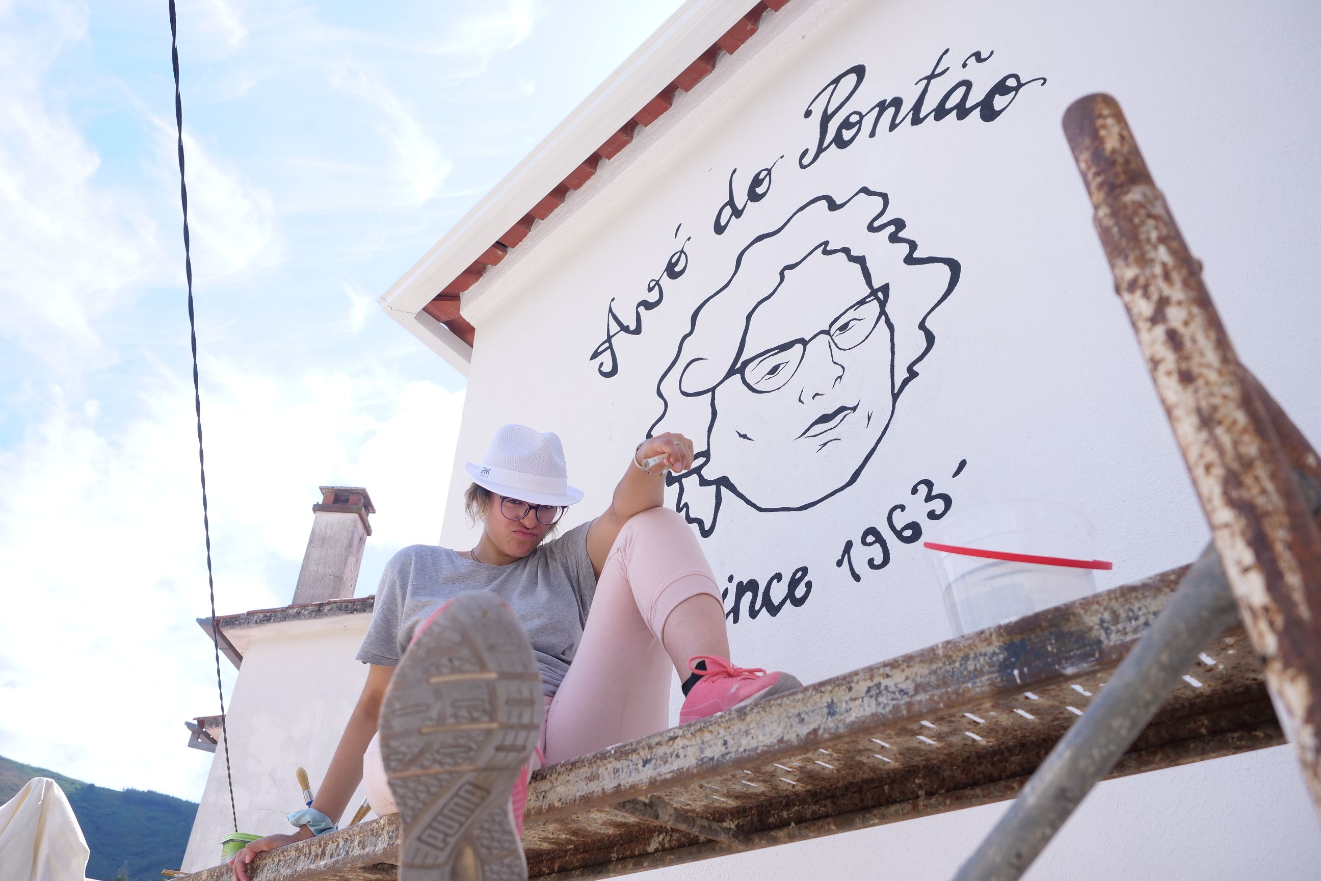 Uma rapariga está sentada em cima de um andaime à frente de um mural a posar para a câmara